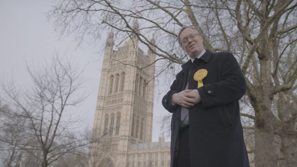 A man in a coat with a yellow rosette stands in front of victoria tower, westminster, looking towards the camera.