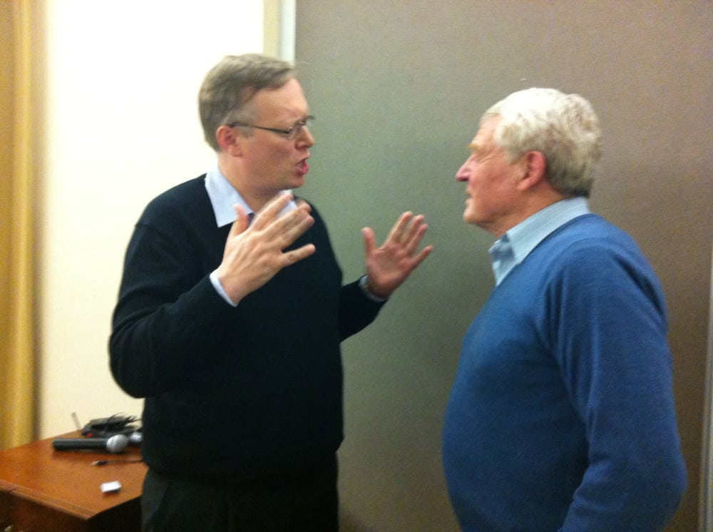 Two men engaging in a conversation, one gesturing with his hands while the other listens attentively, in a room with a beige wall.