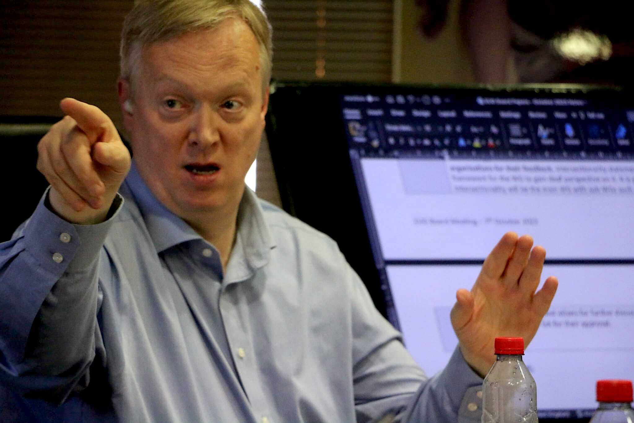 A man in a blue shirt pointing and speaking in a meeting room with two water bottles on the table and a computer screen displaying notes in the background.