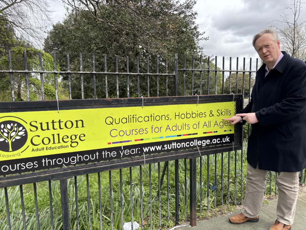 A middle-aged man pointing at a sutton college sign that advertises qualifications, hobbies, and skills courses for adults of all ages.