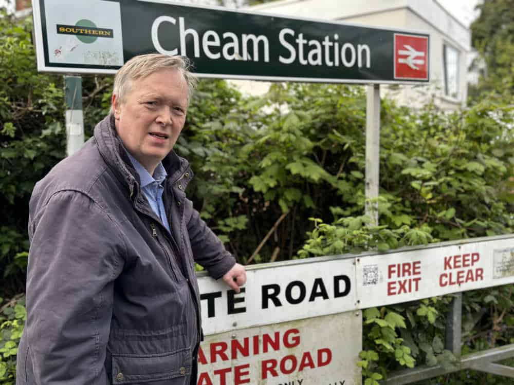 A man in a jacket standing beside the sign of cheam station, looking at the camera.