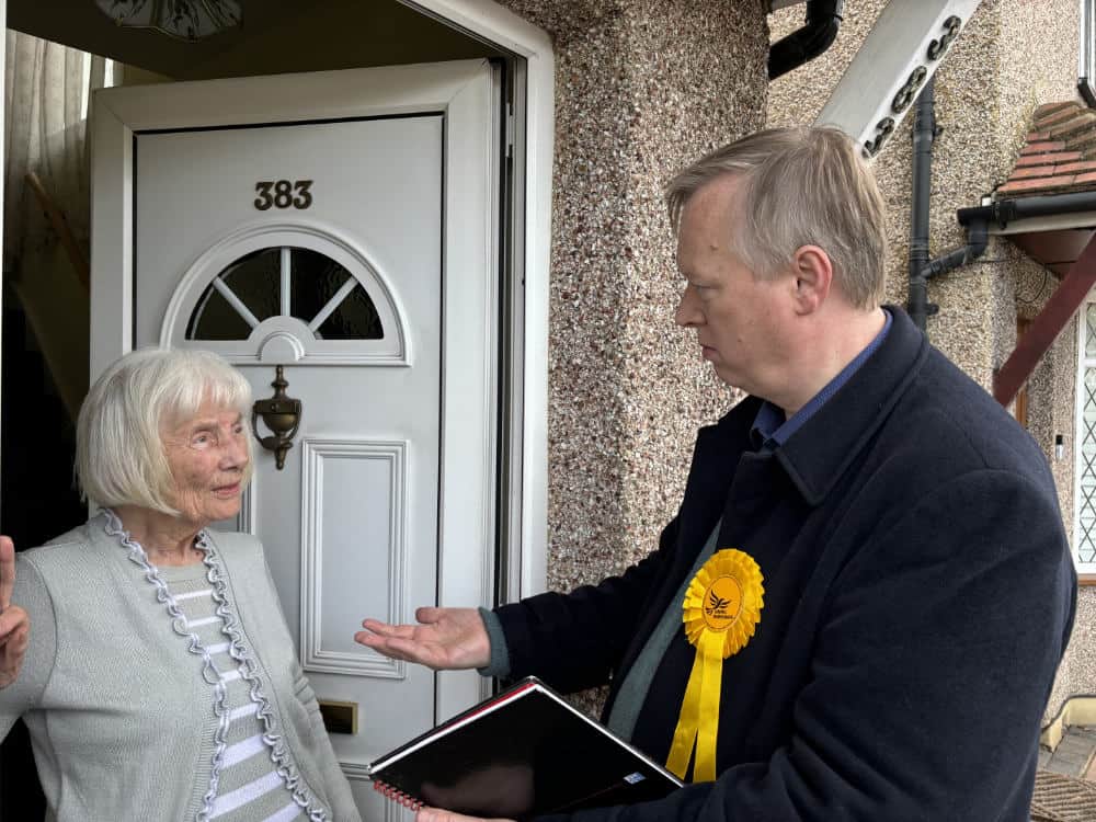 A man with a rosette, holding a clipboard, talks to an elderly woman at her front door.