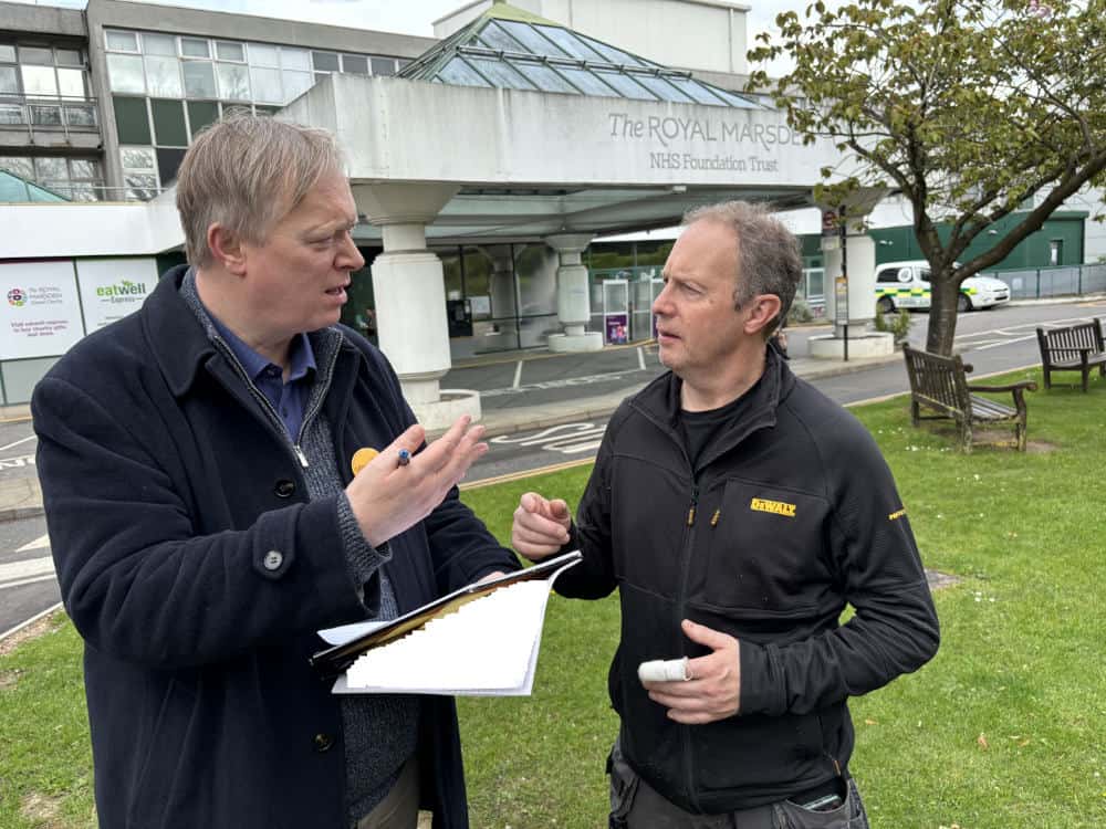 Two men having a discussion outside the royal marsden hospital; one holds a clipboard and the other a bottle.