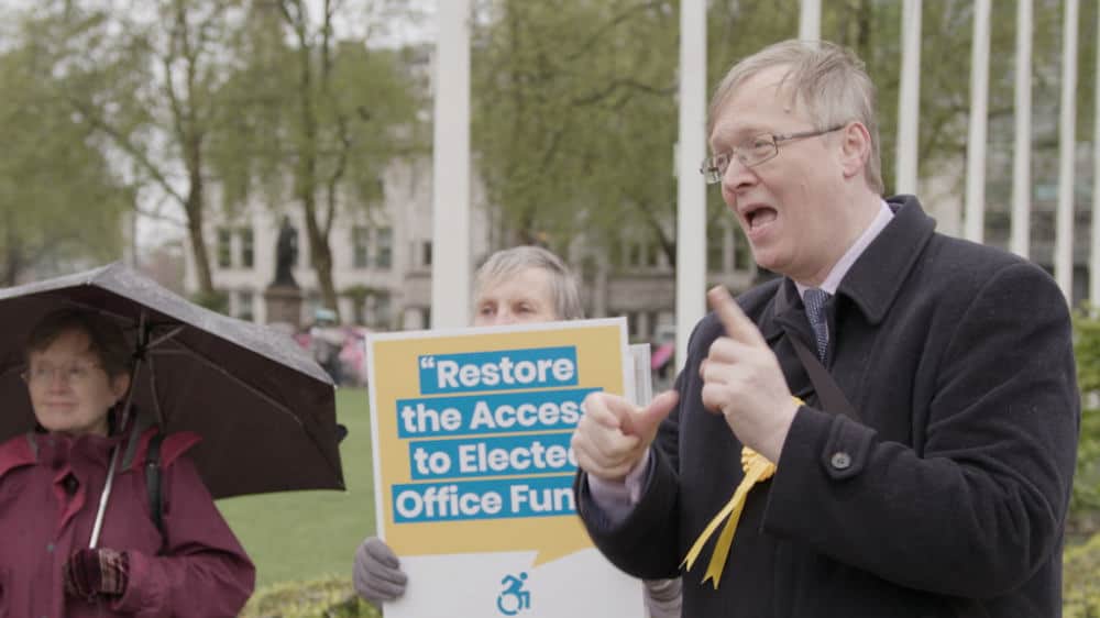 Man speaking passionately at an outdoor rally, holding a sign that says "restore the access to electoral office fund." other people visible in the background.