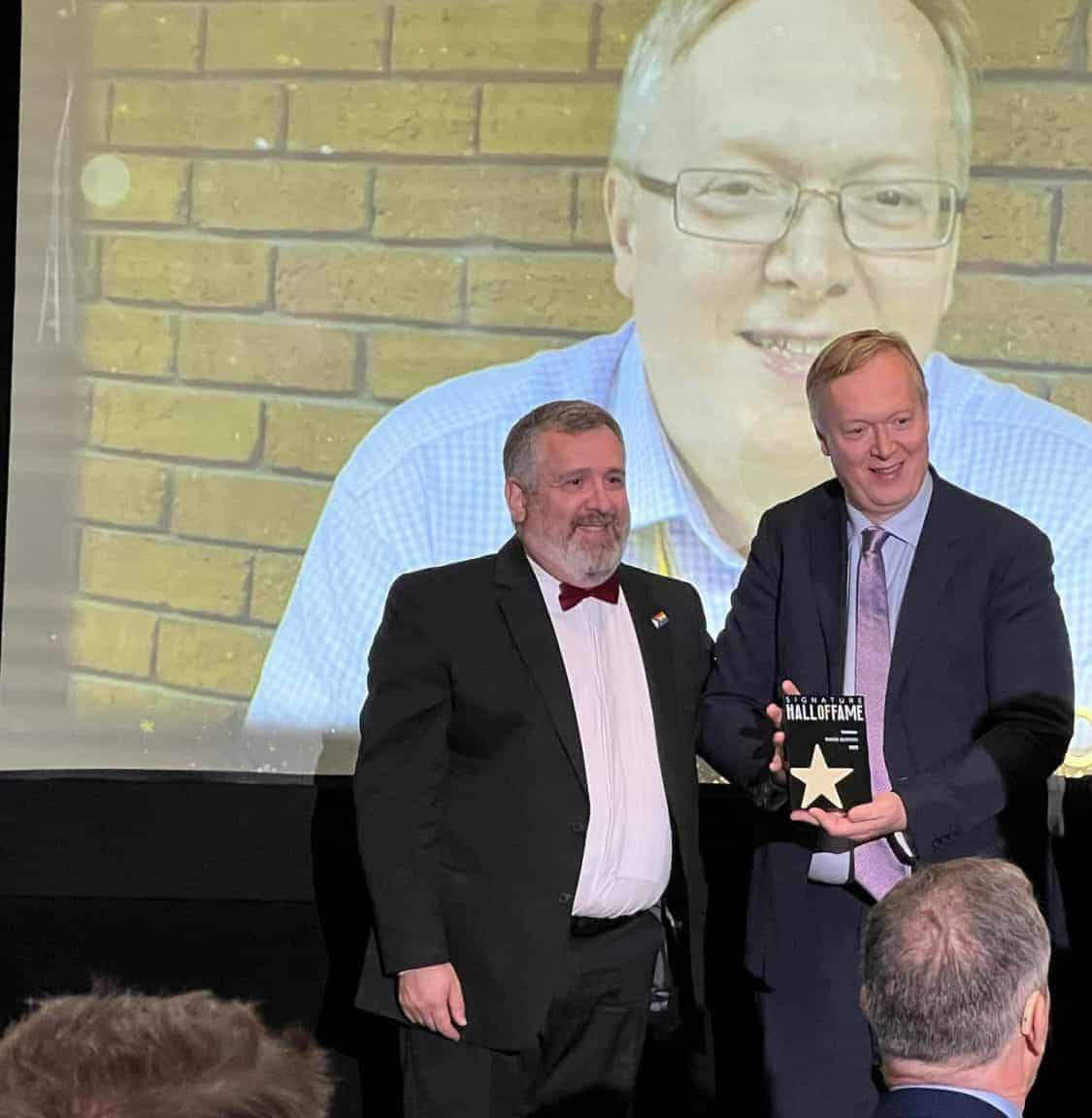 Two men, one holding an award, standing in front of a screen displaying a smiling man's image at an indoor event.