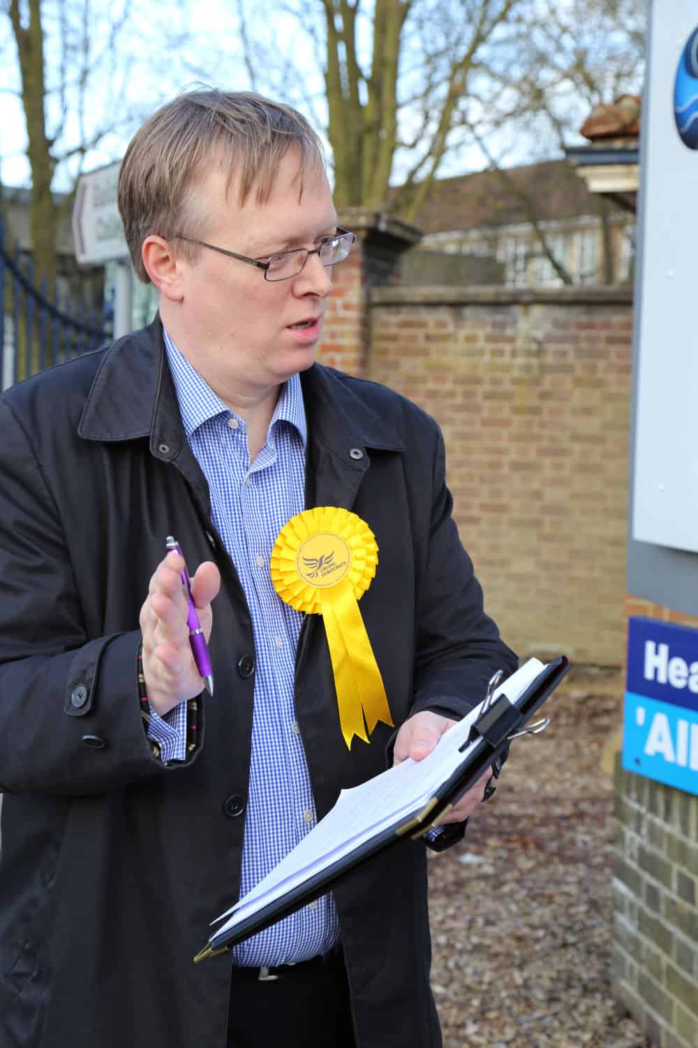 Man in a coat with a yellow ribbon badge holding a clipboard and pen, speaking outdoors near a signpost.