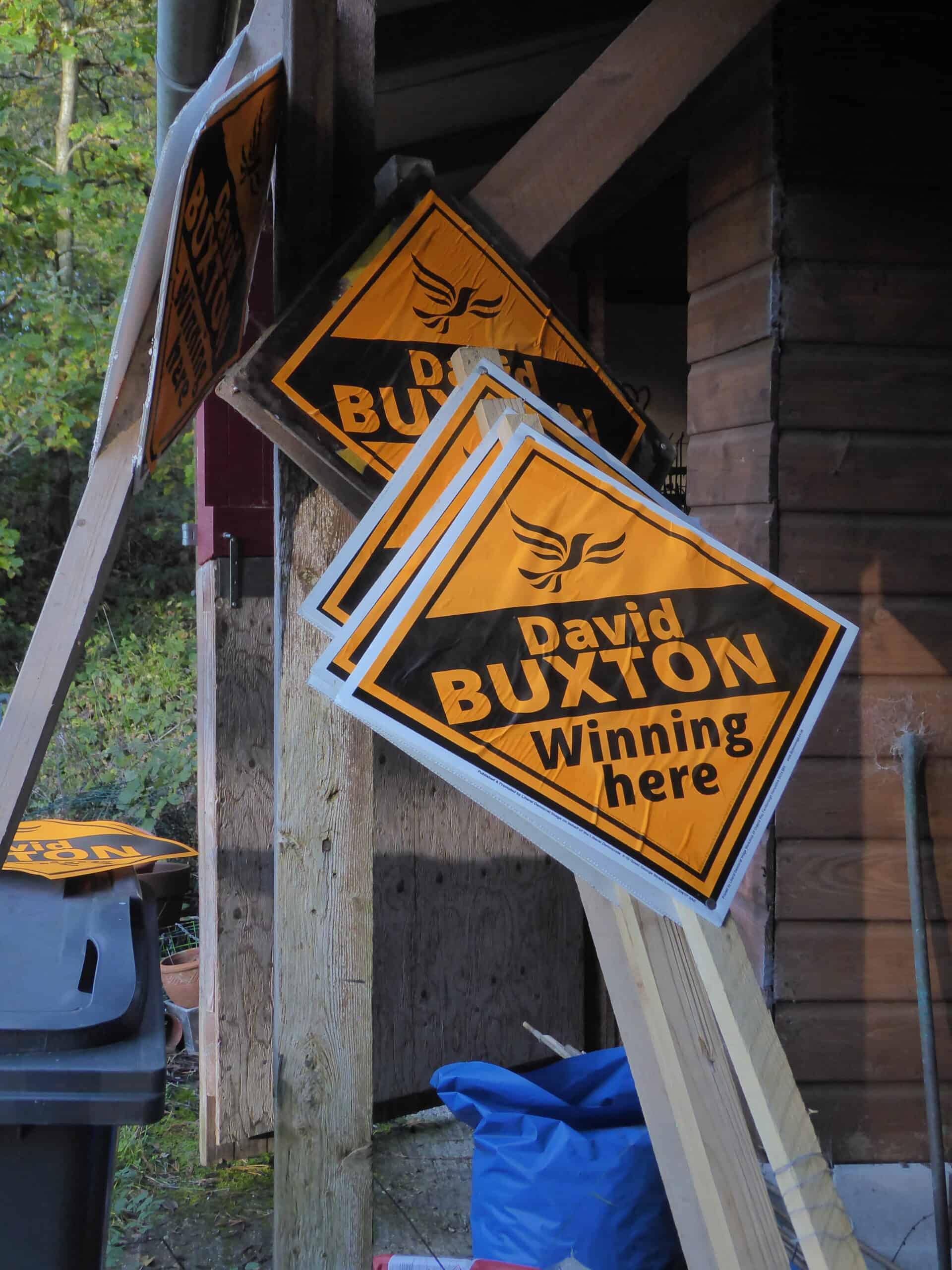 Stack of yellow campaign signs for david buxton leaning against a wooden structure in a wooded area.