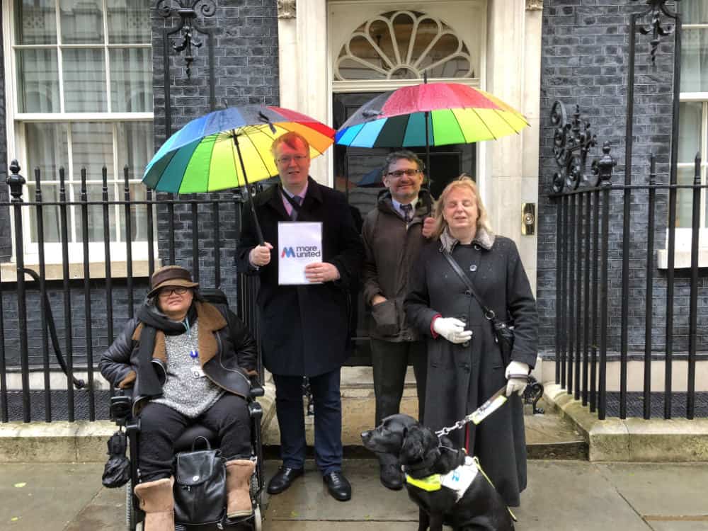 Four people with colourful umbrellas and two guide dogs standing in front of a black ornate gate, holding a sign that says "more united.
