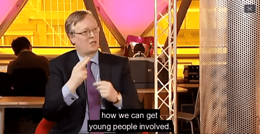Man in glasses and suit gestures while speaking in a studio with pink lighting in the background. subtitles about involving young people are visible.