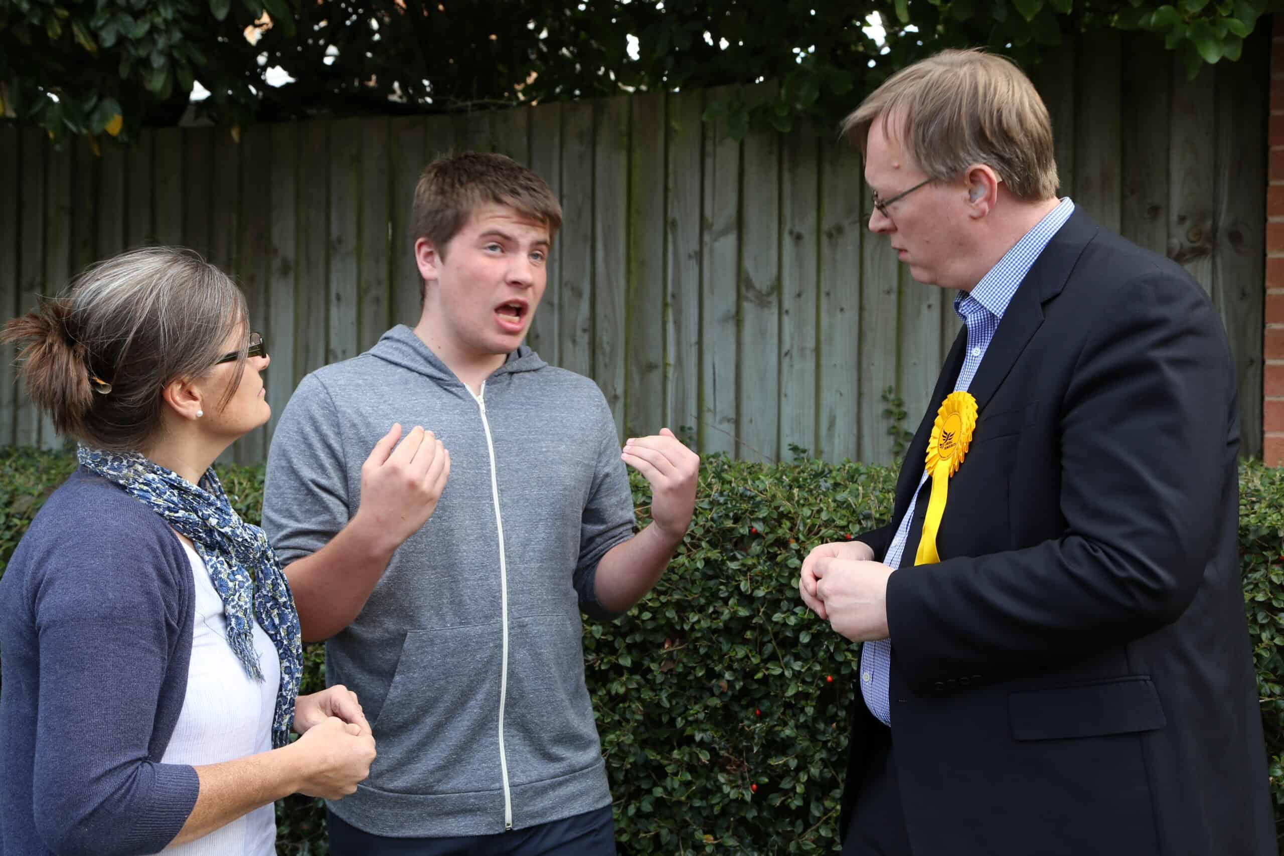 Two men and a woman engaged in a discussion outdoors, one man wearing a yellow rosette.