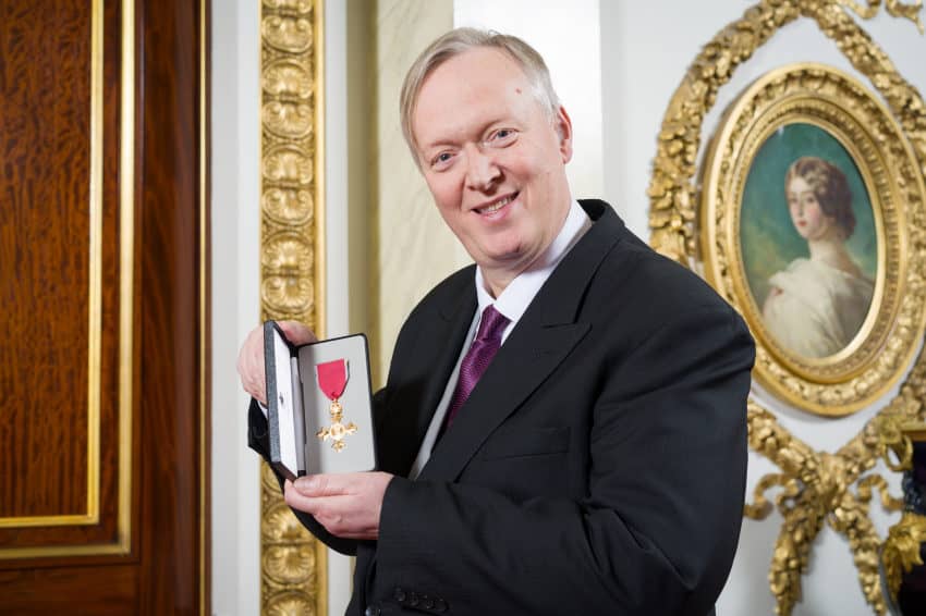 Man in suit smiling at the camera while holding an award with a red ribbon inside an elegant room with a portrait on the wall.
