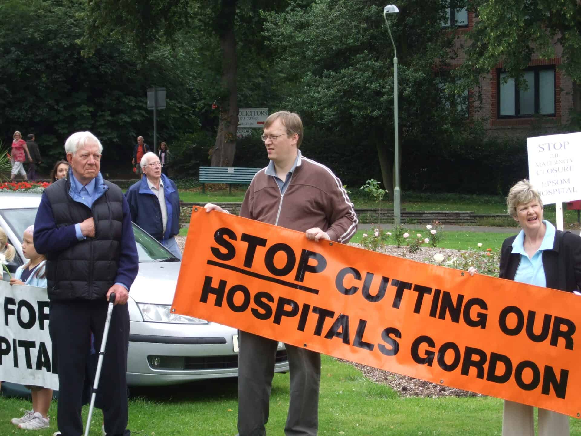 Group of people demonstrating with a banner reading "stop cutting hospitals gordon" in a park-like setting.