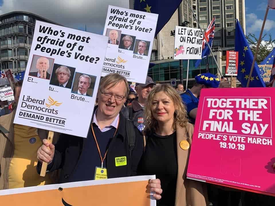 Two individuals proudly holding signs at the people's vote march, expressing support for a final say on brexit, on a sunny day with a crowd in the background.