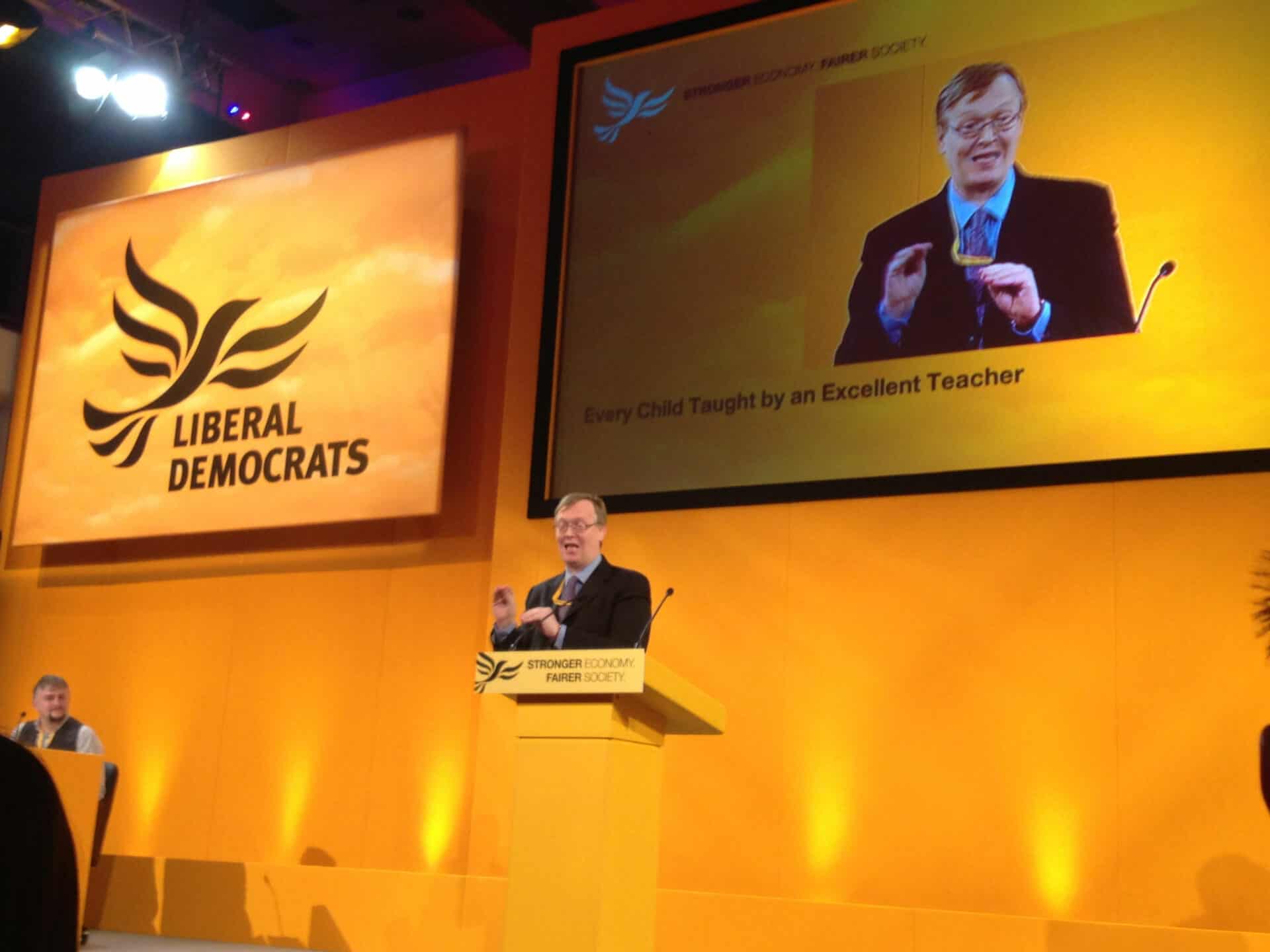 A man speaks at a podium with a liberal democrats logo, with a large screen behind him displaying the same scene and text about education.