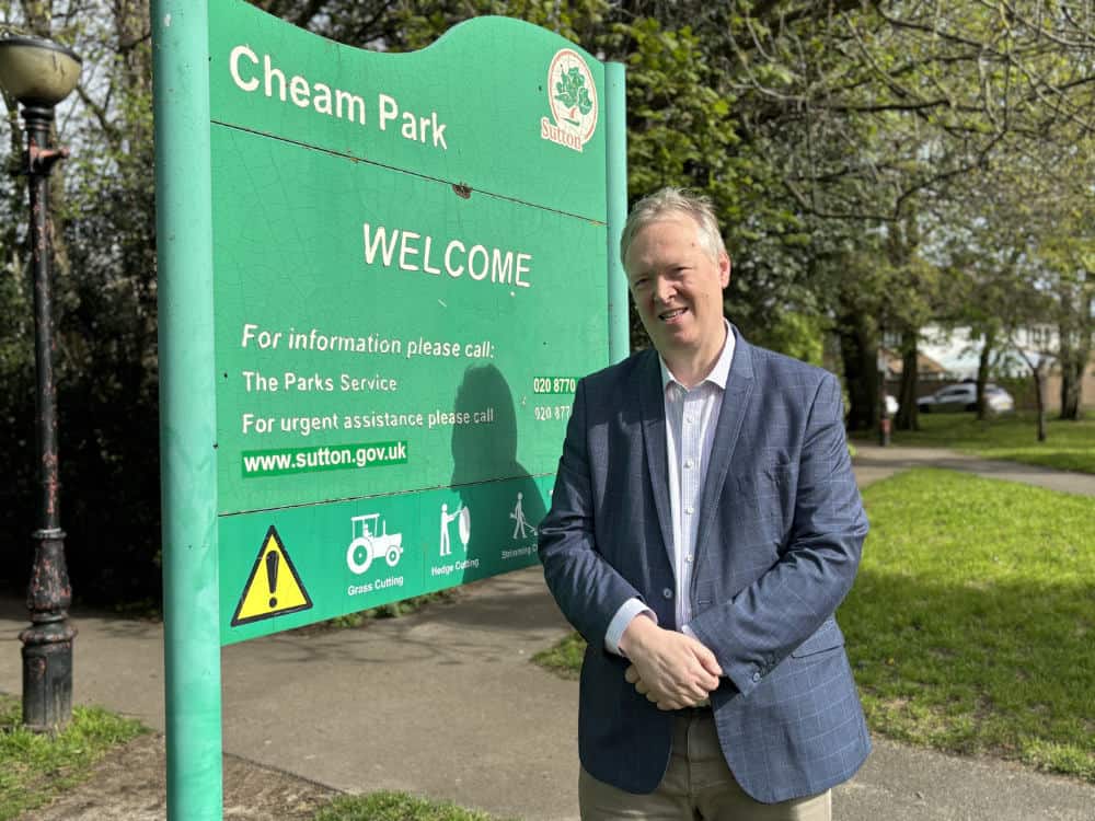 A man standing beside a "welcome to cheam park" sign, smiling on a sunny day.