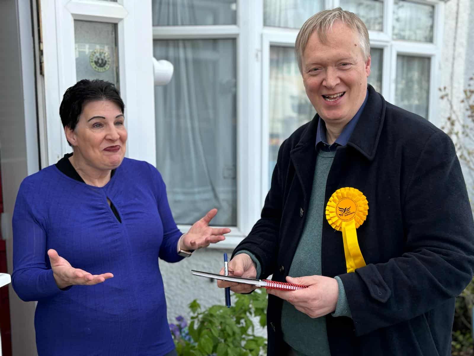 A woman gestures animatedly while talking to a smiling man with a yellow rosette, who holds a clipboard, outside a house.