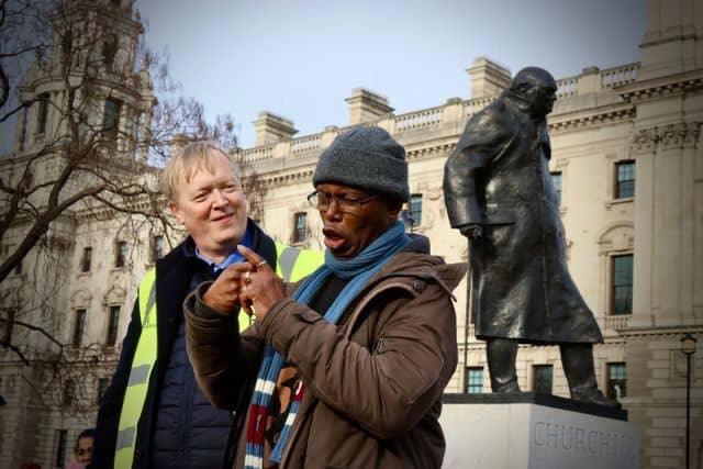Two men having a discussion outdoors with a statue of winston churchill in the background. one wears a hi-vis vest, and the other is wearing a hat and glasses.